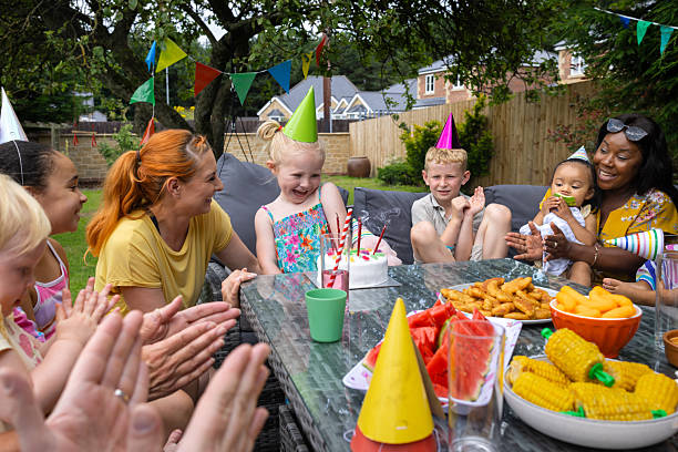 A wide shot of a cheerful group of young children and parents sitting together outdoors, enjoying birthday celebrations on a sunny day. They are enjoying a fun summer birthday garden party together with family and friends. The young girl is smiling on her birthday, and on the table are party food and snacks.

Videos are available similar to this scenario.
