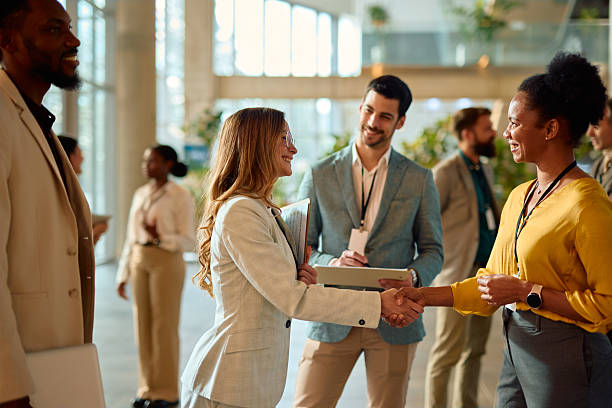 Two businesswomen shaking hands during a conference meeting, engaging in networking while other professionals converse in the background, highlighting collaboration and partnership